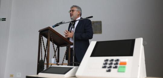 Fotografia de um homem falando em pé em uma tribuna. Ele veste um terno e atrás dele há uma pare...