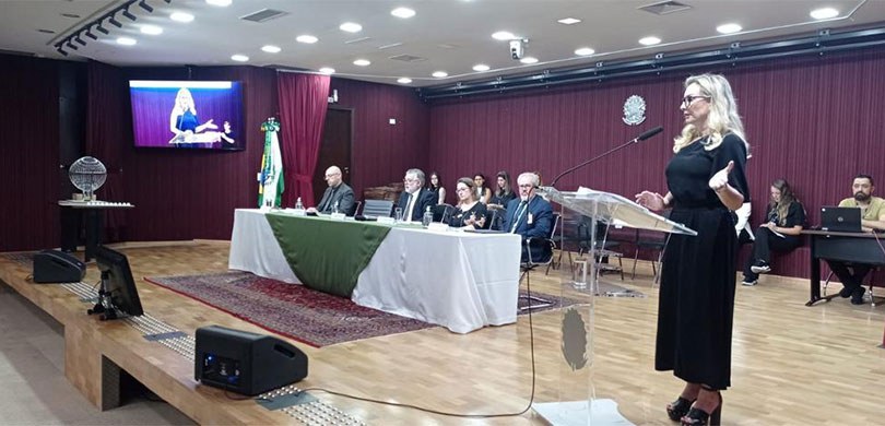 Fotografia do auditório do TRE-PR. No palco, é possível ver a mesa de autoridades e uma mulher f...