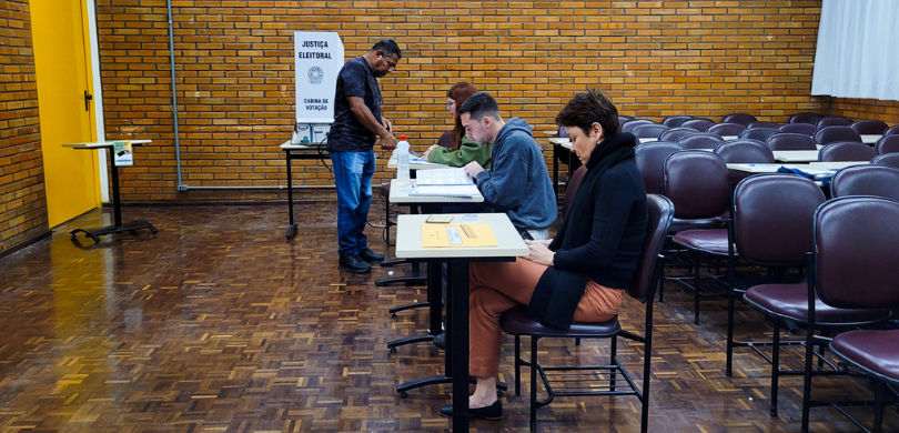 Fotografia em que se observa um ambiente interno de votação com quatro pessoas. No centro da ima...