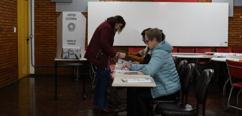 Fotografia em que se observam quatro pessoas em um ambiente interno de votação. Em primeiro plan...