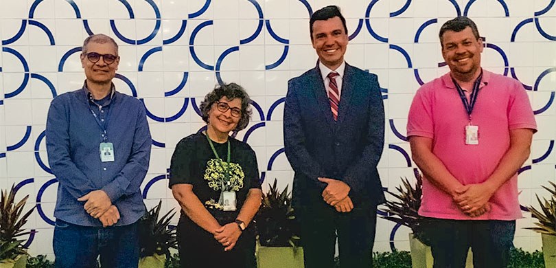 Fotografia de quatro pessoas, três homens e uma mulher, posando sorrindo para foto com as mãos c...