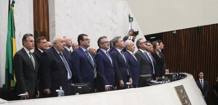 Fotografia de diversos homens e duas mulheres de perfil atrás de uma bancada branca. Todos veste...