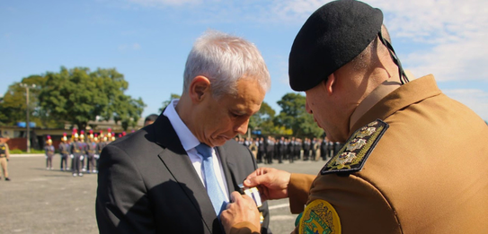 Na imagem, observa-se um militar de perfil fixa a referida medalha na lapela do homem de terno. ...