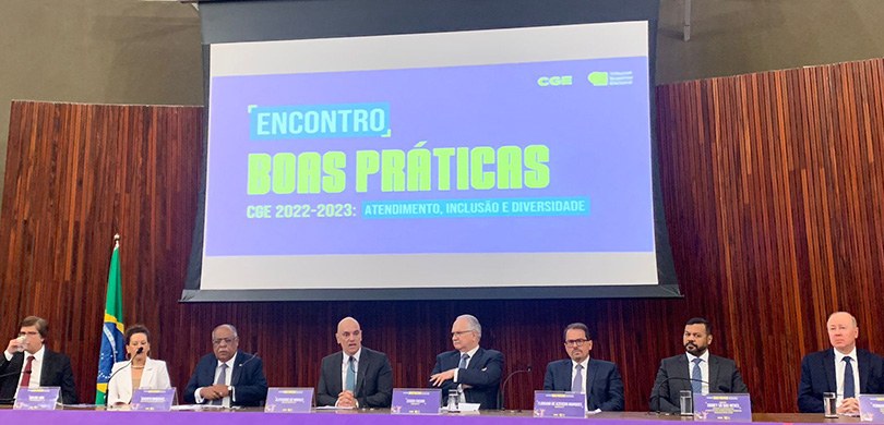 Fotografia de uma mesa de autoridades com sete homens e uma mulher. Ao fundo, há um telão em que...