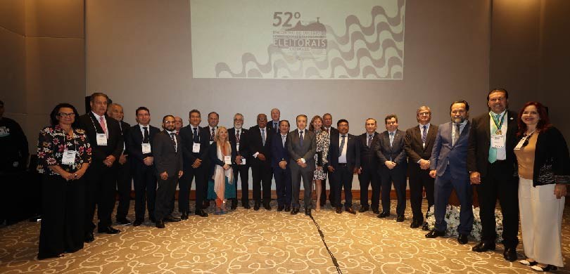 Fotografia de várias pessoas, organizadas em um meio círculo, posando sorrindo em frente a um te...
