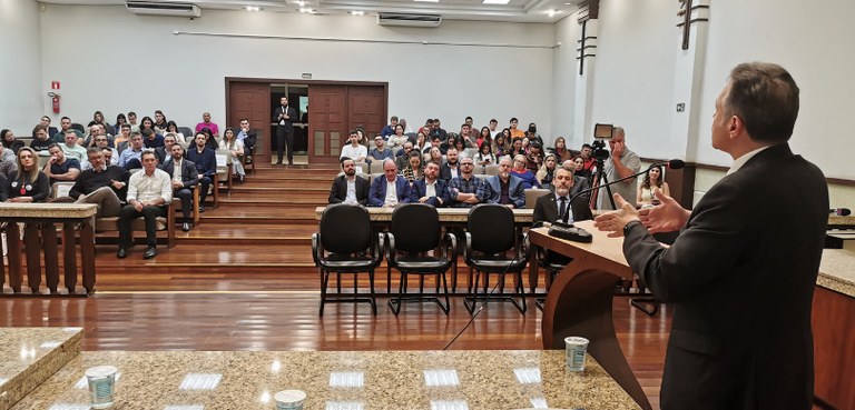 Fotografia do desembargador Panza posicionado atrás de um púlpito de madeira durante palestra. E...