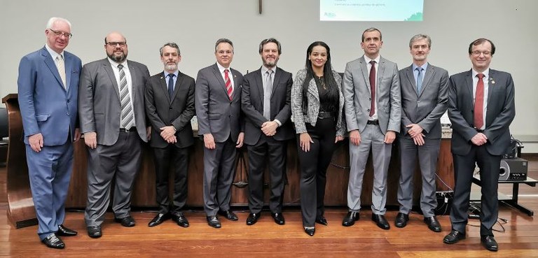 Fotografia de oito homens de terno e uma mulher, que veste calça e camisas pretas e um blazer ci...
