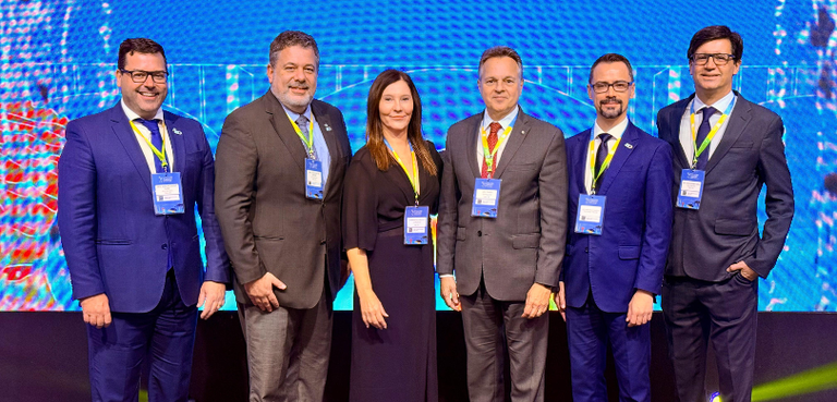 Fotografia de cinco homens de terno e uma mulher, que usa vestido preto, posando sorrindo em fre...