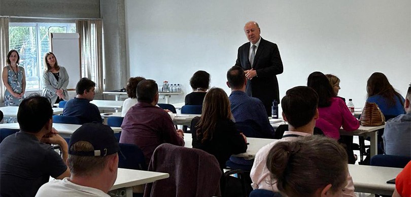 Fotografia de um homem de terno falando diante de homens e mulheres sentados em carteiras escola...