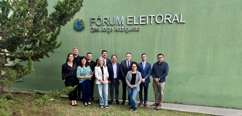 Fotografia de quatro mulheres e sete homens posando sorrindo em frente ao Fórum Eleitoral Des. J...