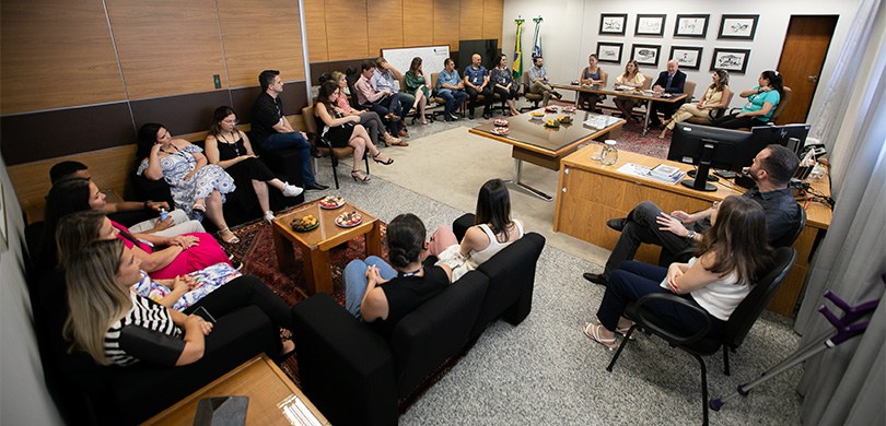 Fotografia de várias pessoas, entre homens e mulheres, reunidas em uma sala, sentadas em cadeira...