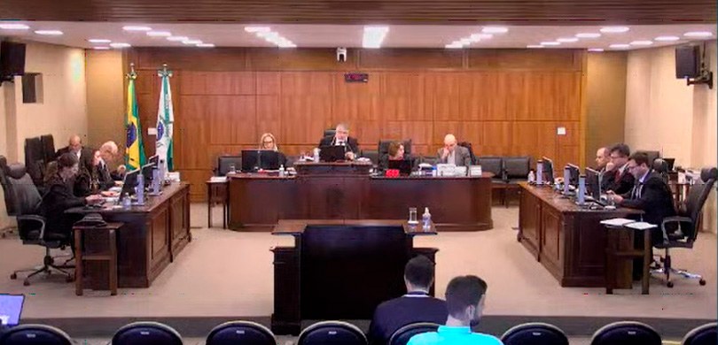 Fotografia da Corte do TRE-PR, reunida na sala de sessões. Ao fundo, há uma bandeira do Brasil e...