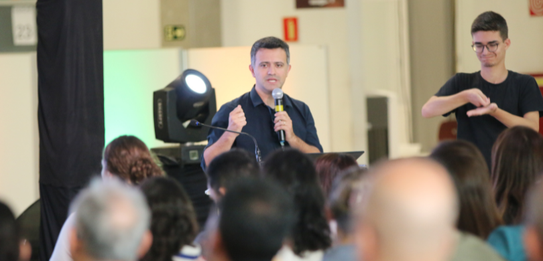Fotografia de nove pessoas, entre homens e mulheres, posando sorrindo em frente a um edifício co...