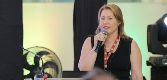 Fotografia de nove pessoas, entre homens e mulheres, posando sorrindo em frente a um edifício co...