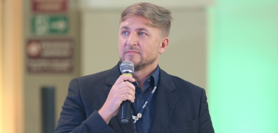Fotografia de nove pessoas, entre homens e mulheres, posando sorrindo em frente a um edifício co...