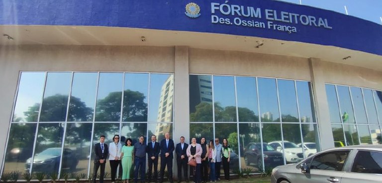 Fotografia de doze pessoas, entre homens e mulheres, posando sorrindo em frente ao Fórum Eleitor...