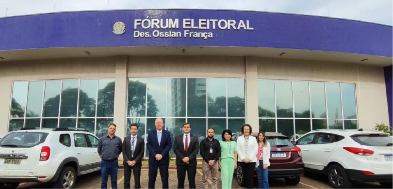 Fotografia de oito pessoas, entre homens e mulheres, posando sorrindo em frente ao Fórum Eleitor...