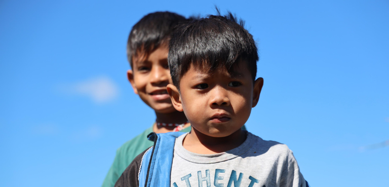 Fotografia de duas crianças indígenas. Ao fundo, há um céu azul.