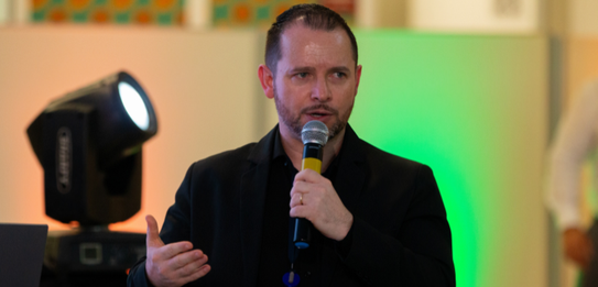Fotografia de nove pessoas, entre homens e mulheres, posando sorrindo em frente a um edifício co...