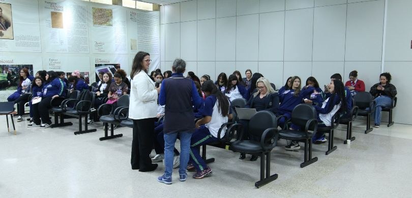 Fotografia na qual é possível ver duas mulheres, em pé, e um grupo de meninas, sentadas, interag...