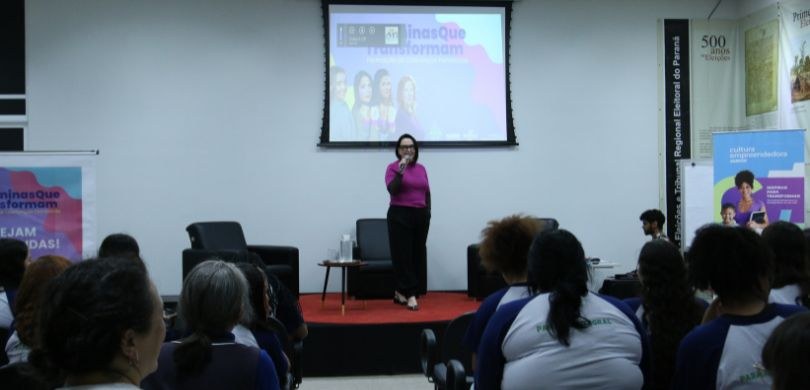 Fotografia da desembargadora eleitoral Tatiane de Cássia Viese em um palco palestrando para as e...