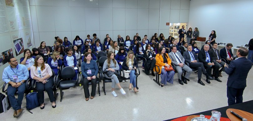 Fotografia tirada de um palco em que aparecem várias estudantes sentadas usando uniformes azuis ...