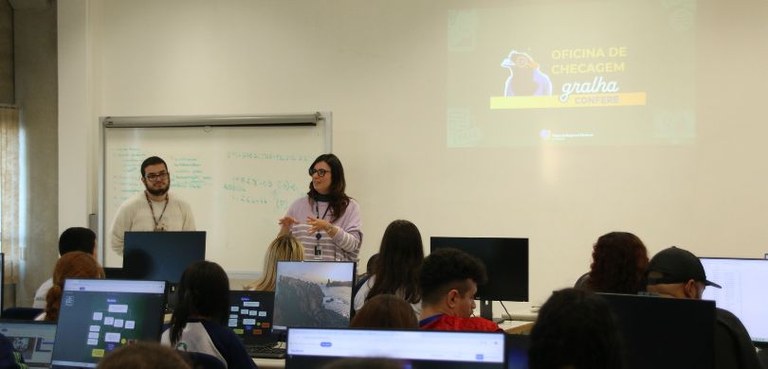 Fotografia em que são vistas em primeiro plano algumas pessoas sentadas em frente a computadores...