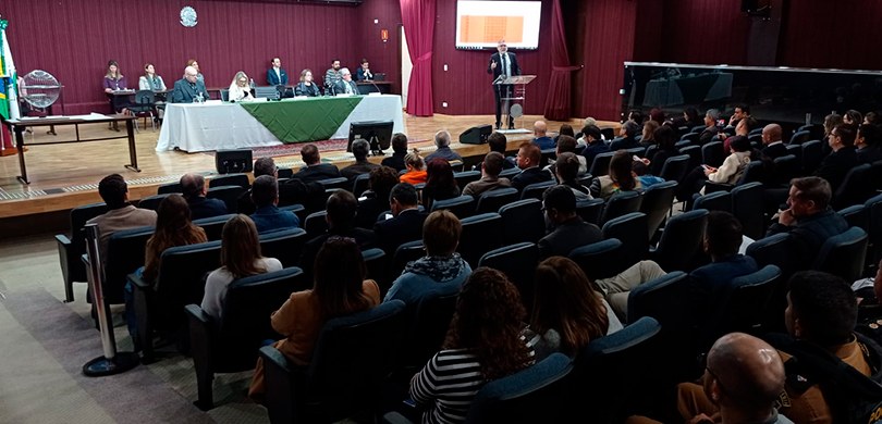 Fotografia do auditório do TRE-PR. No palco, é possível ver a mesa de autoridades e um homem fal...