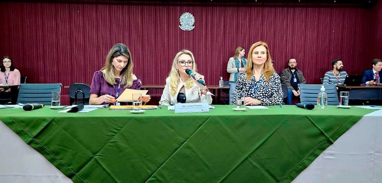 Fotografia do palco de um auditório. A mesa de autoridades é composta por três mulheres, uma del...