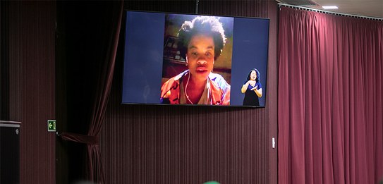 A fotografia mostra uma televisão e atrás um painel e uma cortina da cor vermelha. A tevê mostra...