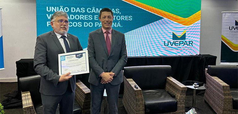 Fotografia de dois homens posando sorrindo em frente a um telão. Um deles segura um certificado....