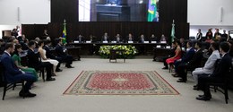 Fotografia de um auditório, com uma mesa de autoridades ao centro. Dos dois lados, há poltronas ...