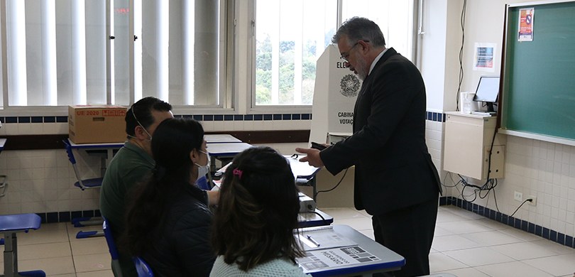Fotografia de um homem em uma seção eleitoral. Ele veste terno e apresenta documentos para as me...