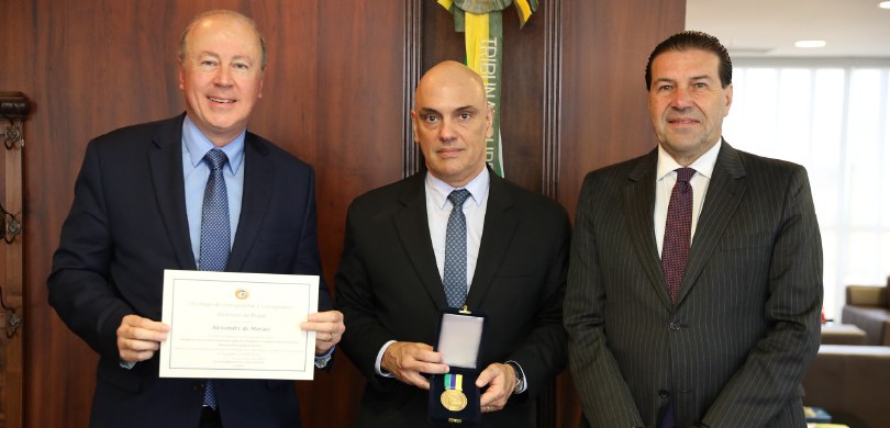 Fotografia de três homens de terno posando sorrindo. O homem ao centro segura uma medalha e o ho...