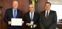 Fotografia de três homens de terno posando sorrindo. O homem ao centro segura uma medalha e o ho...