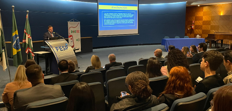 Fotografia em que se observa um auditório durante uma palestra. À esquerda, em plano médio, um h...
