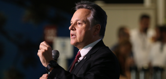 Fotografia de nove pessoas, entre homens e mulheres, posando sorrindo em frente a um edifício co...