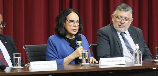A fotografia mostra uma televisão e atrás um painel e uma cortina da cor vermelha. A tevê mostra...