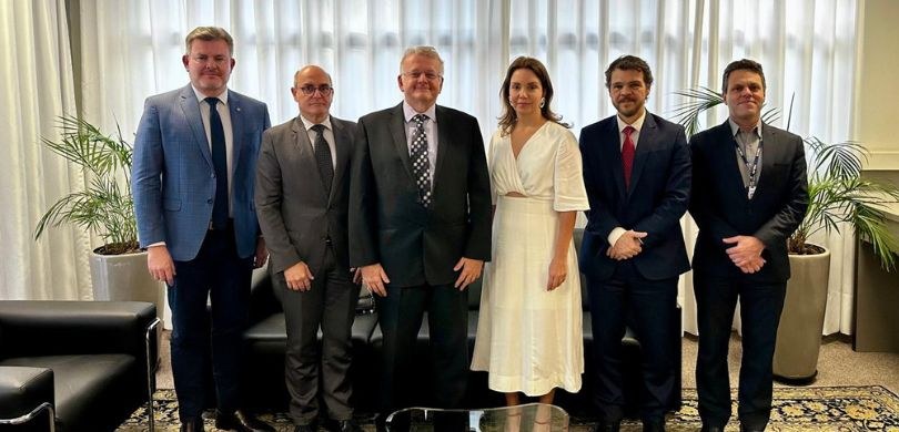 Cinco homens e uma mulher posam para a foto em uma sala com carpete claro, cortinas brancas e so...