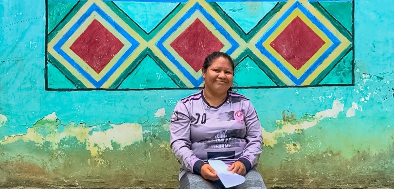 Fotografia em que se observa uma mulher indígena, sentada e sorrindo levemente para a câmera. El...