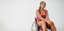 Fotografia de uma jovem mulher negra em uma cadeira de rodas. Ela veste uma camisa estampada e c...