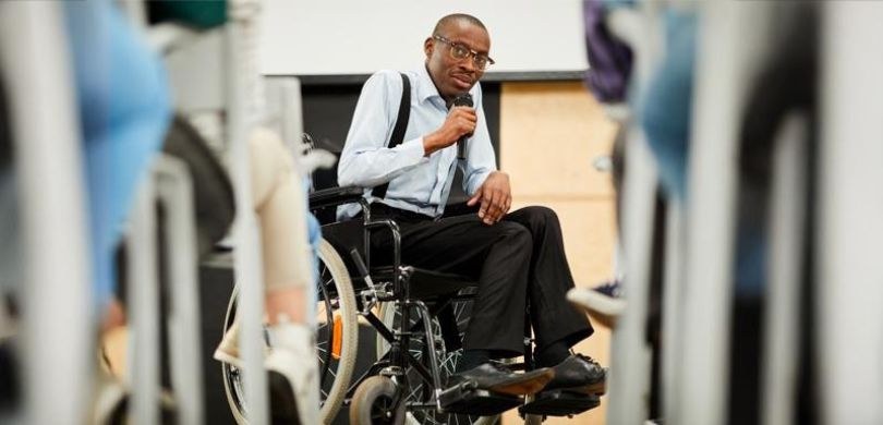 Na imagem, vê-se um homem negro de óculos de grau, sentado em uma cadeira de rodas. Ele veste um...