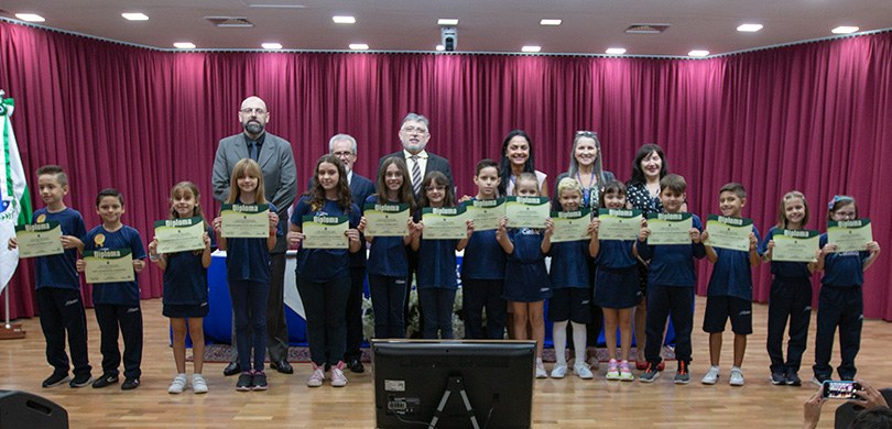 Fotografia de 15 crianças, três homens de terno e três mulheres posando sorrindo para foto. As c...