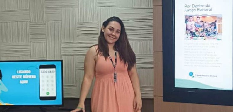 Uma mulher de cabelos escuros, usando vestido na cor pêssego, posa para a foto. Ao seu lado esqu...