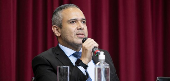 A fotografia mostra uma televisão e atrás um painel e uma cortina da cor vermelha. A tevê mostra...