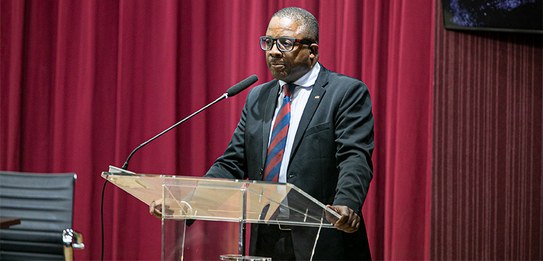 A fotografia mostra uma televisão e atrás um painel e uma cortina da cor vermelha. A tevê mostra...