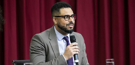 A fotografia mostra uma televisão e atrás um painel e uma cortina da cor vermelha. A tevê mostra...