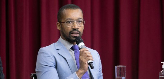 A fotografia mostra uma televisão e atrás um painel e uma cortina da cor vermelha. A tevê mostra...