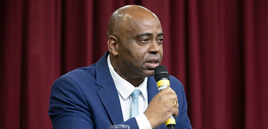 A fotografia mostra uma televisão e atrás um painel e uma cortina da cor vermelha. A tevê mostra...