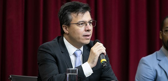 A fotografia mostra uma televisão e atrás um painel e uma cortina da cor vermelha. A tevê mostra...